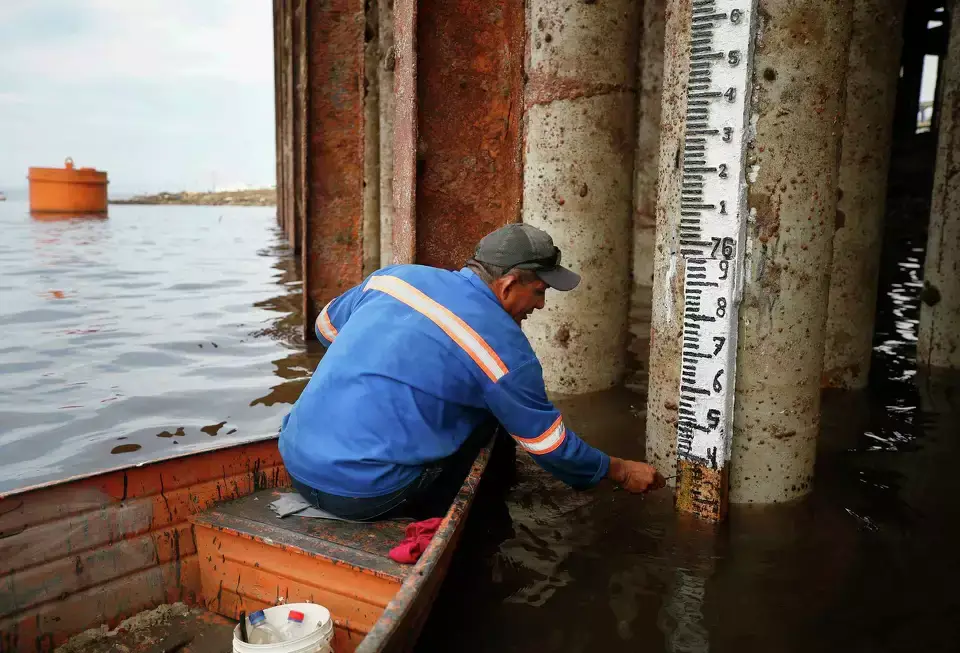 A dock worker in a life vest stands in shallow water, reading a meter stick to measure the critically low water level of the Negro River at Manaus port.