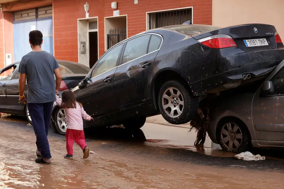 Individuals walk beside damaged vehicles on a street following severe flooding in Guadassuar, Valencia region, Spain.