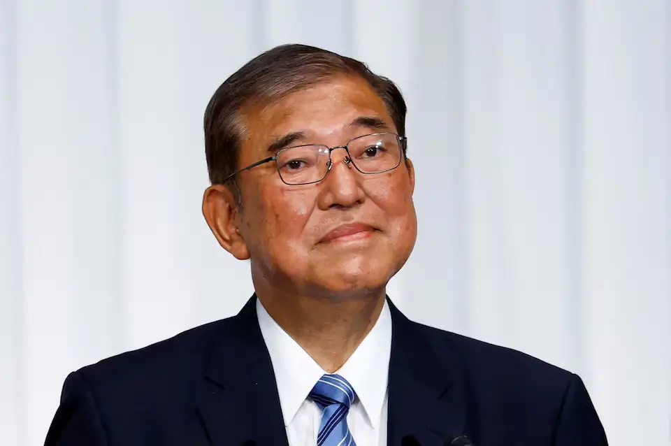 Prime Minister Shigeru Ishiba walks into the press conference room wearing a dark suit, his face serious as he approaches the podium amidst camera flashes at LDP headquarters.