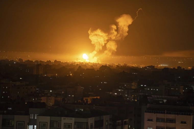 Large explosion illuminates night sky over urban area, with smoke plume rising above silhouetted buildings in foreground.