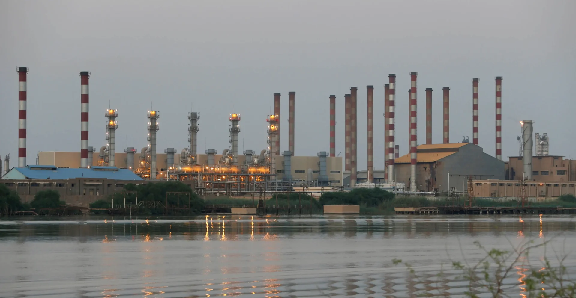 Industrial refinery with smokestacks along a river at dusk, flares burning on either side.