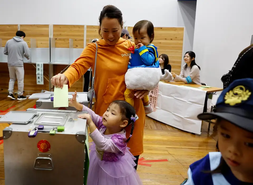 An elderly Japanese woman casting her ballot behind a blue privacy screen while election workers in face masks observe at a local Tokyo polling station.