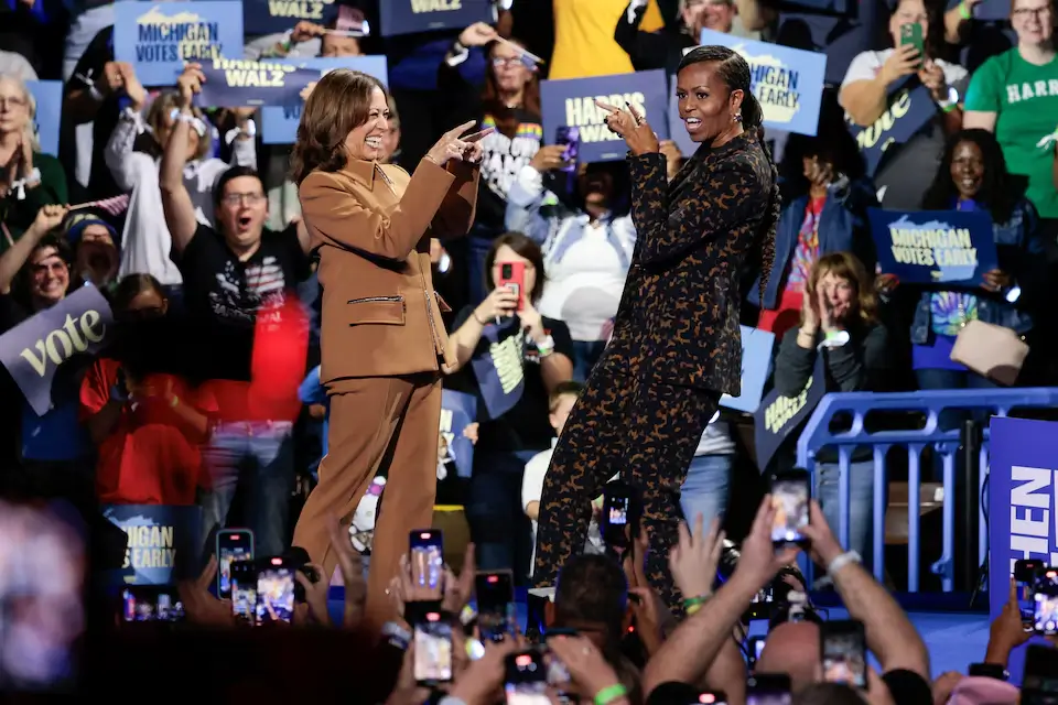 Vice President Kamala Harris and Michelle Obama share an embrace on stage while smiling warmly at a campaign rally in Kalamazoo, their figures silhouetted against bright stage lighting.