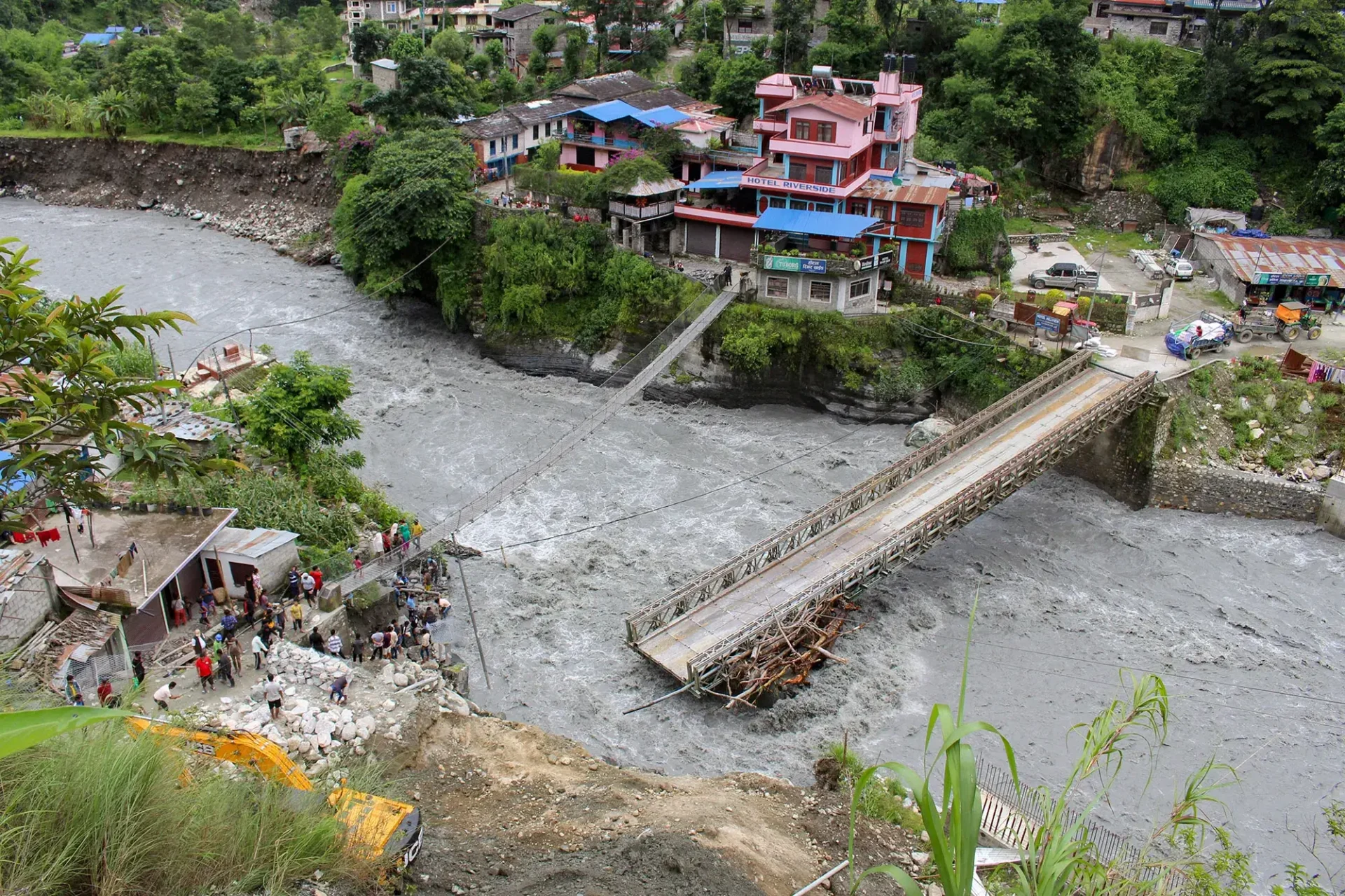 Flooded river in Nepal surges past a damaged bridge. Colorful buildings cling to riverbanks as people gather near swollen waters.