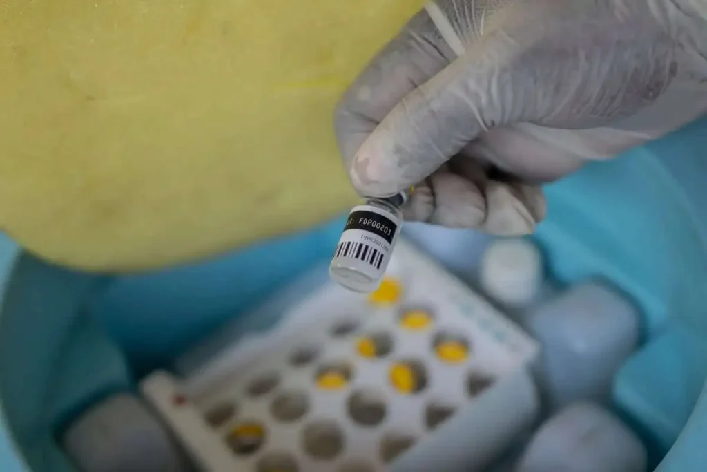 A healthcare worker in medical scrubs holds up a clear glass vial labeled as mpox vaccine against a blurred clinical background.