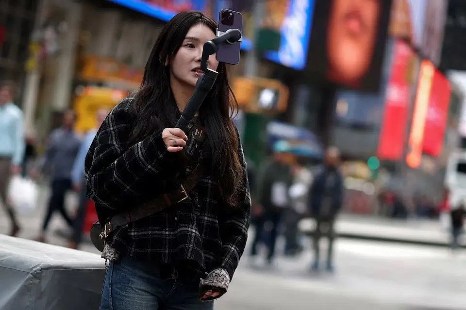 A woman recording a video on her smartphone in front of the iconic Times Square billboards and screens in New York City.