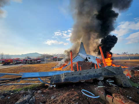 Multiple firefighters in protective gear spray water and foam onto a burning aircraft wreckage, with thick black smoke rising from the crash site at Muan International Airport.