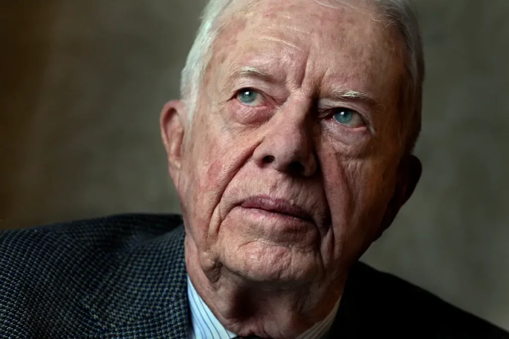 Close-up portrait of Jimmy Carter showing his intense blue eyes and weathered face against a dark background, wearing a textured blazer.