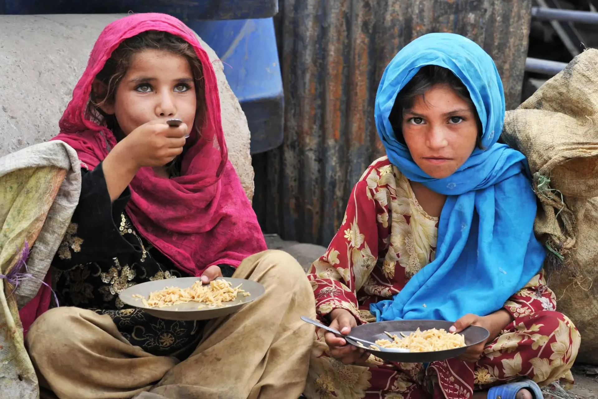 Two Young Afghan girls wearing colorful scarves, sitting on the ground and eating plain food from metal plates.