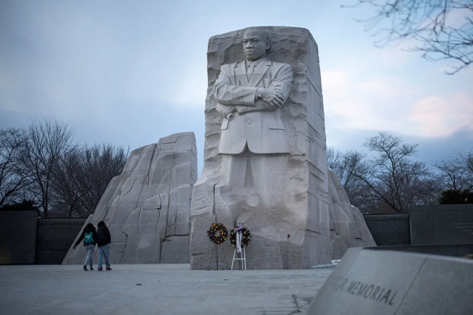 A calm image of tourists passing the imposing granite statue of Martin Luther King Jr. rising from the Stone of Hope on a winter day in Washington, DC, with the memorial framed by cherry blossom trees.